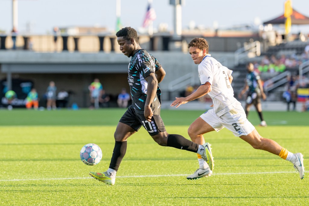 Velocity's Medgy Alexandre dribbles a soccer ball while chased by a Westchester SC defender in a white kit