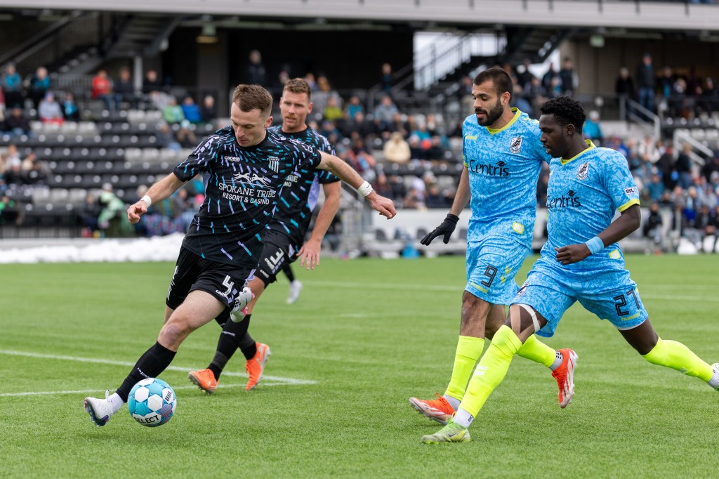 Spokane Velocity defender Simon Fitch dribbles a soccer ball ahead of two Union Omaha players in blue jerseys.