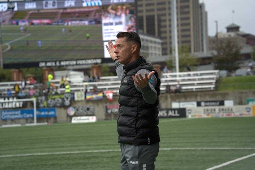 Coach Leigh Veidman, wearing a black vest, raises his arms in disbelief on a soccer field